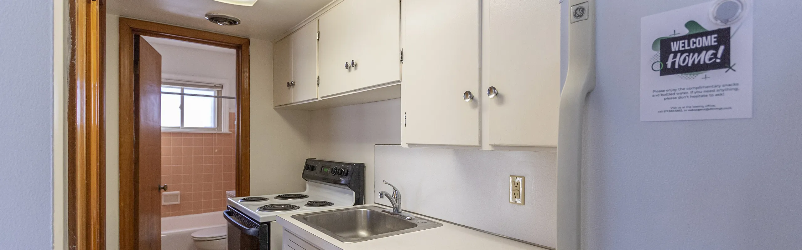 Cozy galley kitchen with bright cabinetry, modern appliances, and warm wood flooring adjacent to a retro-style pink-tiled bathroom.