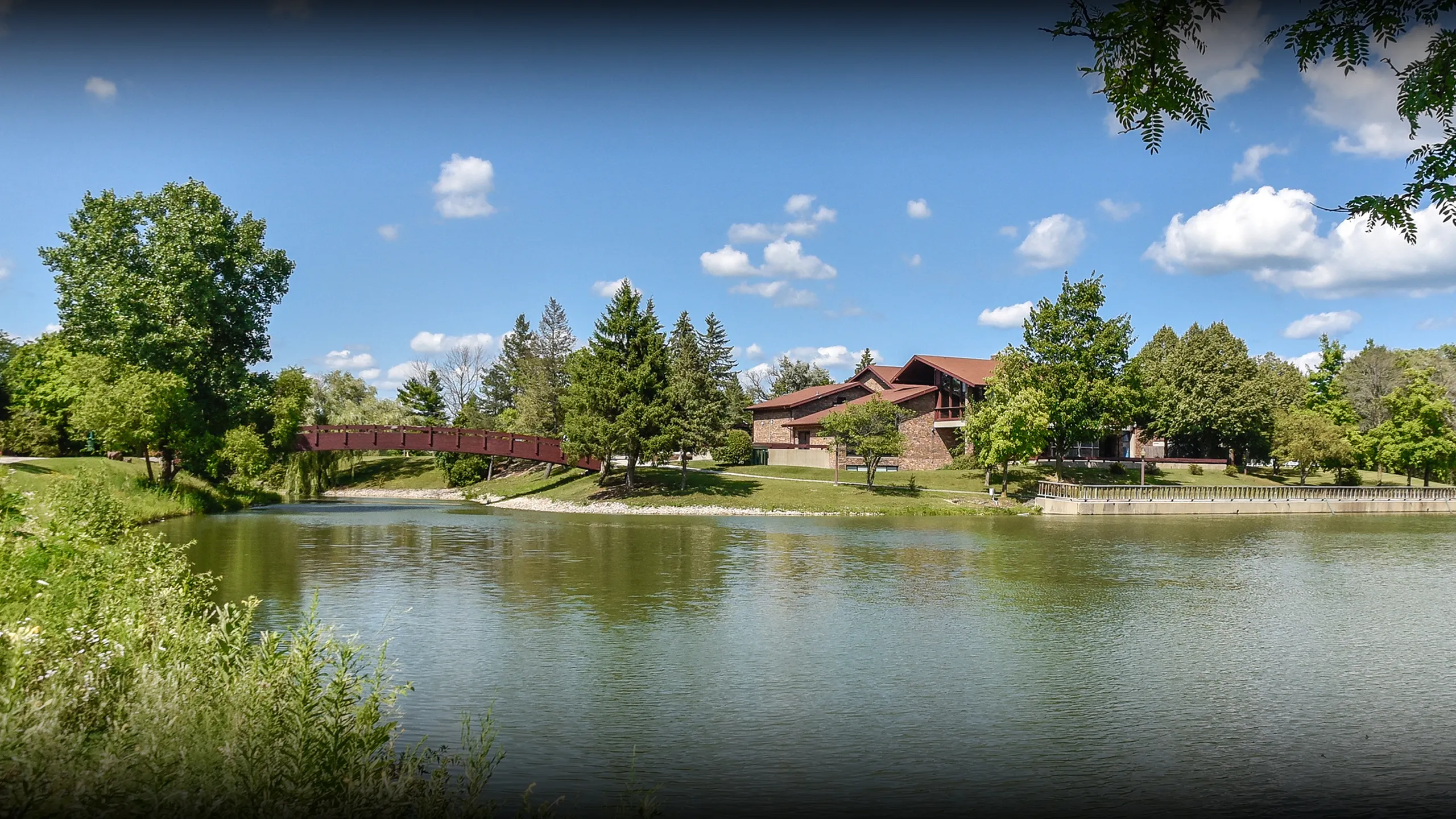 Serene riverside view of a charming brick building surrounded by lush greenery and a scenic bridge on a sunny day.