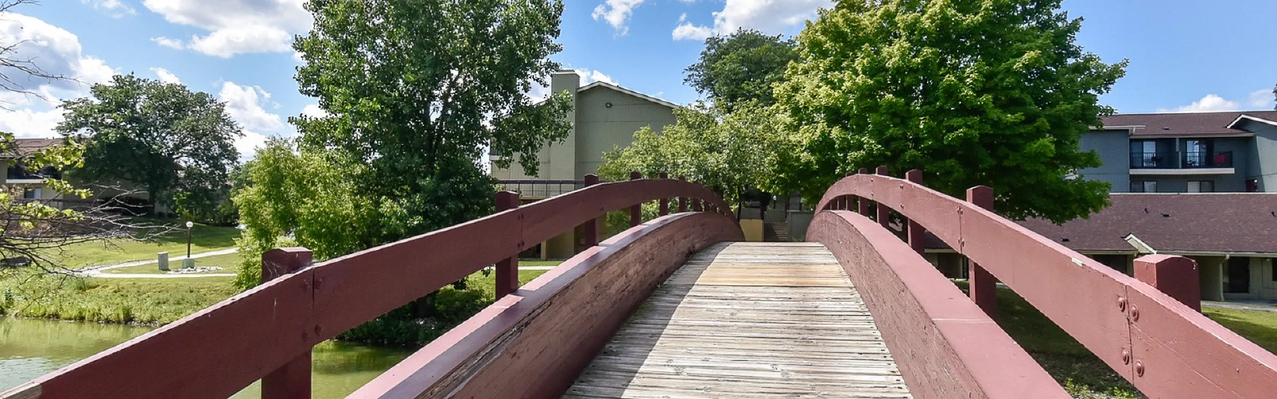 Charming wooden bridge crossing a serene pond, surrounded by lush greenery and modern apartments under a bright blue sky.