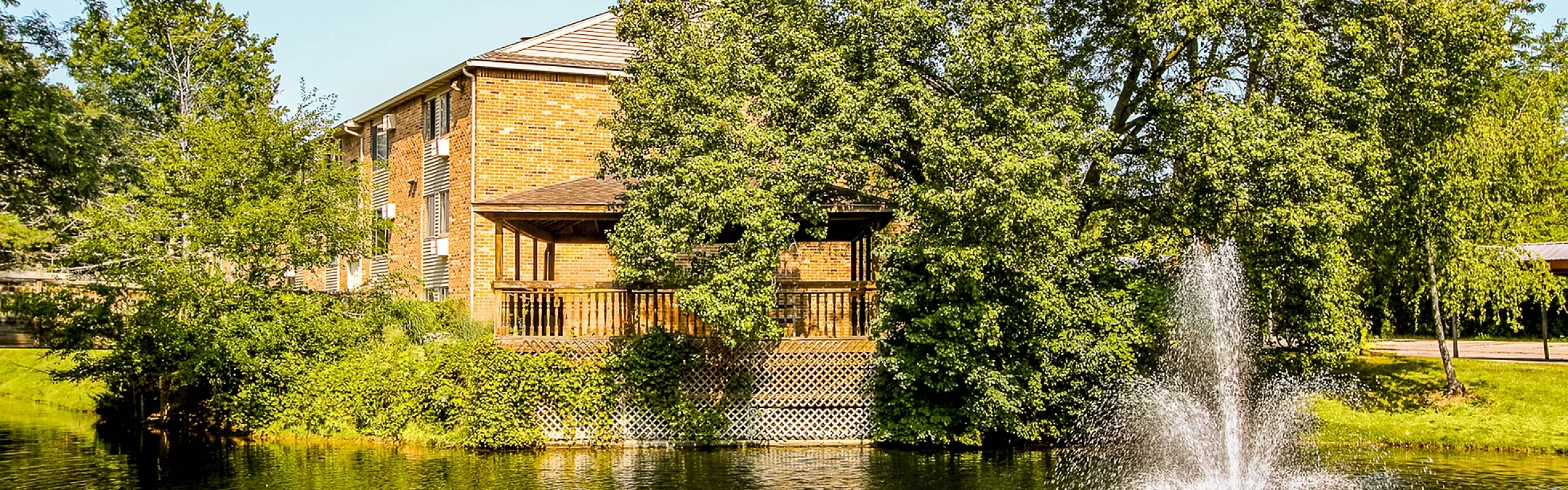 Charming brick apartment building with a scenic view of a tranquil pond and elegant fountain, surrounded by lush greenery.