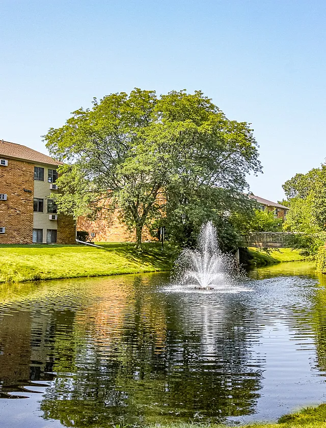 Charming apartment building with a serene pond and fountain, surrounded by lush greenery, offering a peaceful living environment.