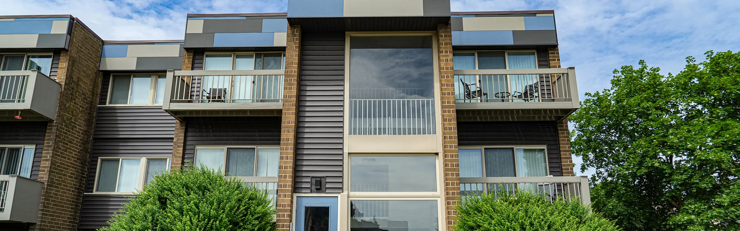 Modern apartment building exterior with stylish geometric design, featuring balconies and lush greenery under a vibrant blue sky.