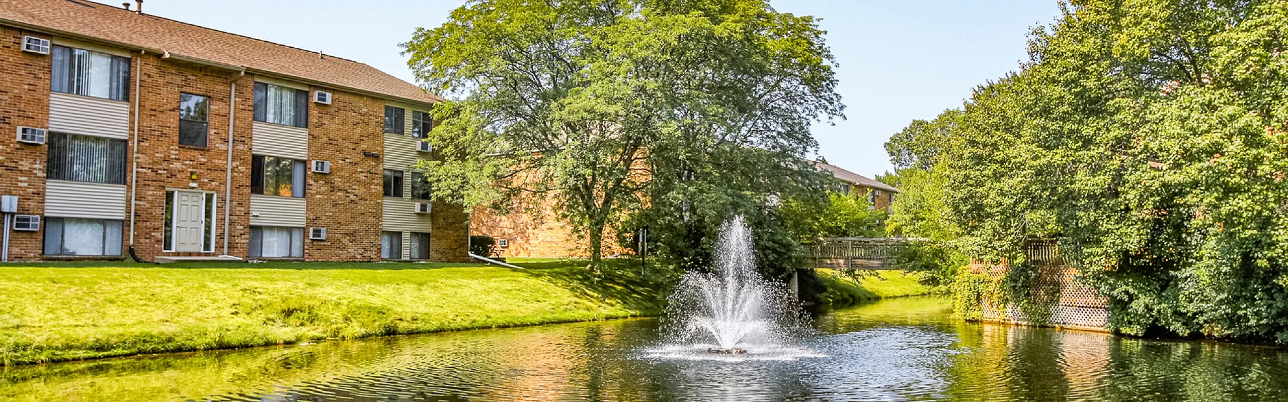 Charming apartment complex with scenic pond and elegant fountain surrounded by lush greenery and sunshine.