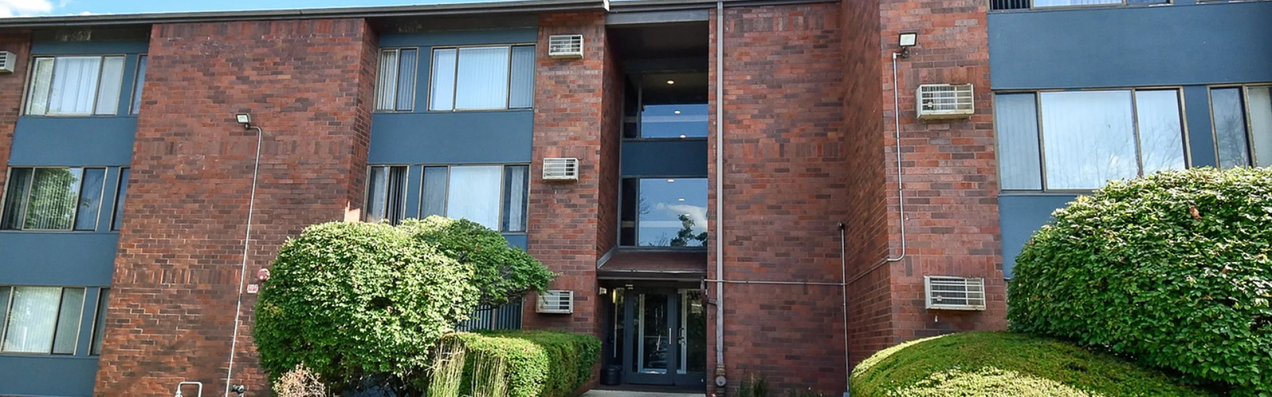 Charming brick apartment building with landscaped greenery and a welcoming entrance under a bright blue sky.