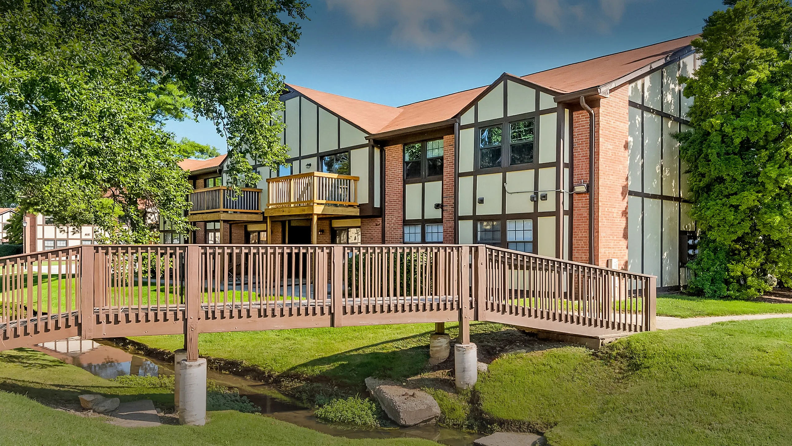 Charming brick and panel apartment exterior with wooden balconies and a scenic footbridge over a lush green lawn.