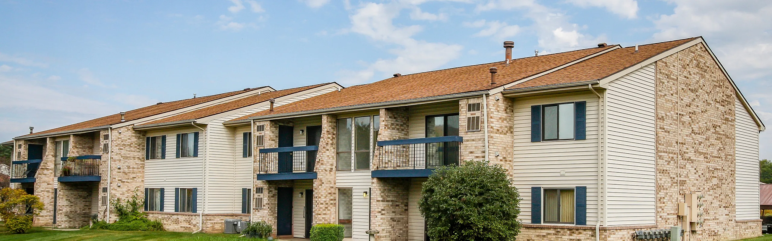 Charming two-story apartment building with beige brick facade, featuring private balconies and lush green surroundings under a bright blue sky.