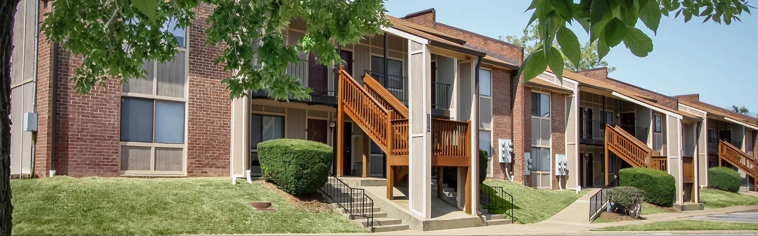Charming red-brick apartment complex with wooden staircases, surrounded by lush greenery and trees, offering a warm and inviting community vibe.