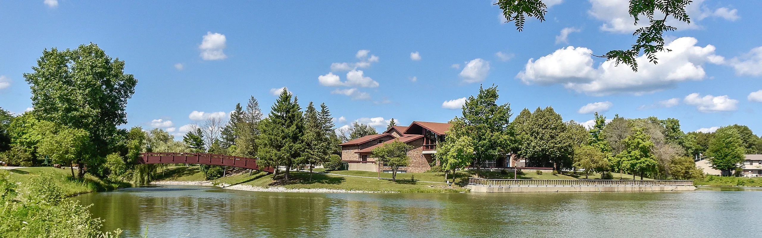 Charming lakeside view featuring a quaint red bridge and lush greenery under a vibrant blue sky.