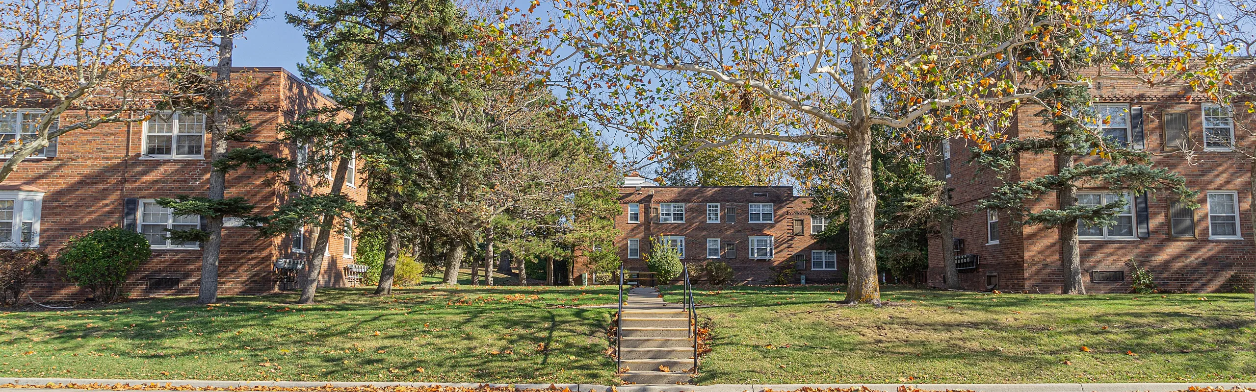 Charming brick apartment complex surrounded by lush green trees and a well-maintained lawn, perfect for serene living.
