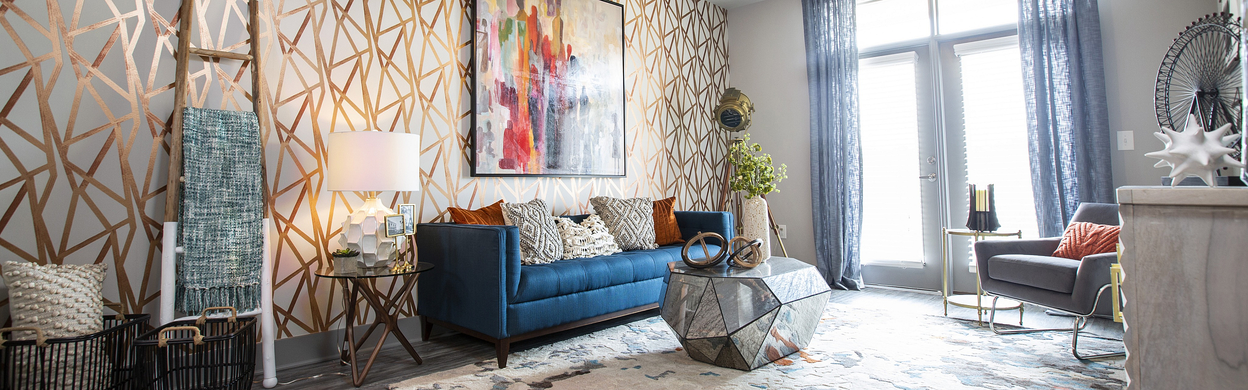 Chic living room with a modern geometric coffee table, vibrant artwork, stylish blue sofa, and elegant gold-accented decor against a patterned feature wall.