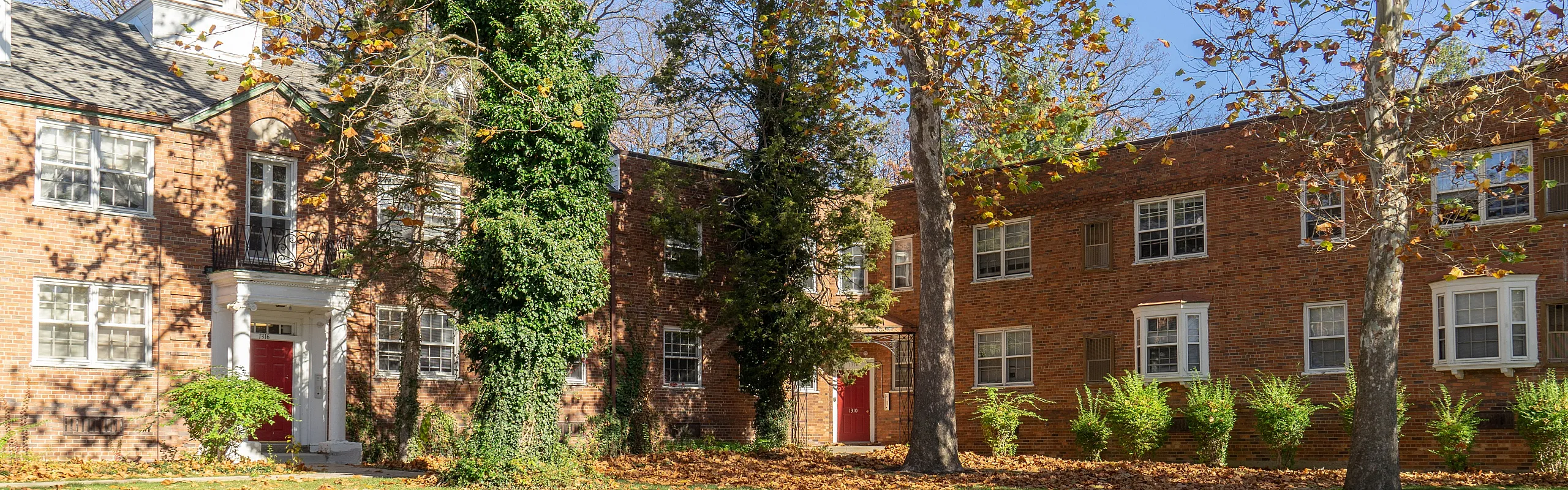 Charming brick apartment building with vibrant red doors, surrounded by lush trees and greenery, capturing a serene and inviting neighborhood vibe.