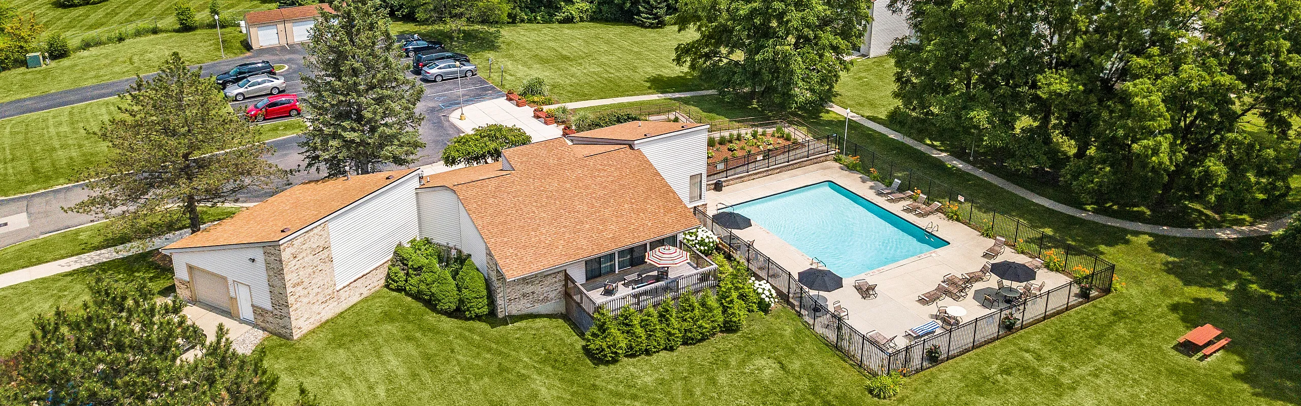 Aerial view of a charming apartment complex with lush green surroundings, featuring a beautiful swimming pool and cozy outdoor seating area.
