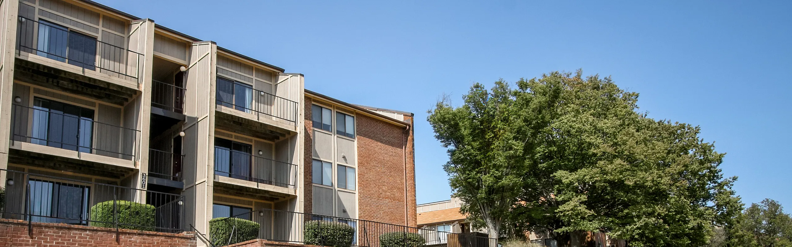 Modern apartment building with spacious balconies set against a vibrant blue sky and surrounded by lush green trees.