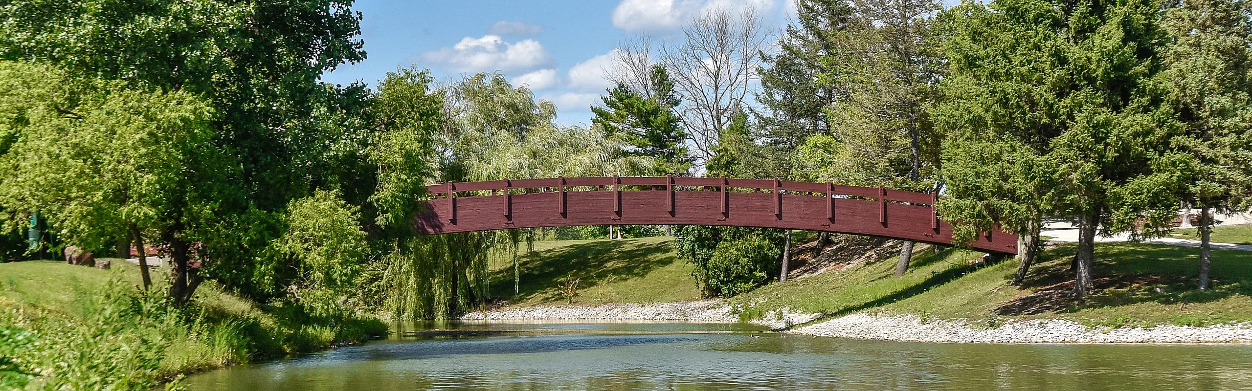 Charming wooden bridge arching over a serene river, surrounded by lush greenery and clear blue skies.
