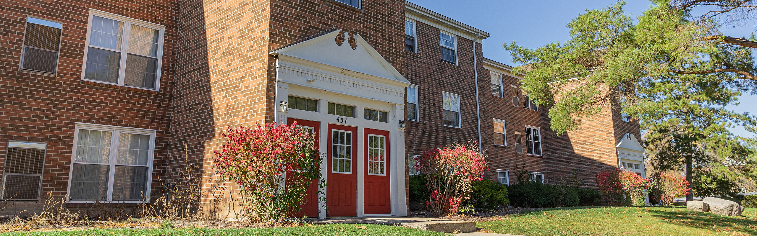 Charming brick apartment building with red doors and manicured landscaping, bathed in warm sunlight, offering a welcoming entrance.