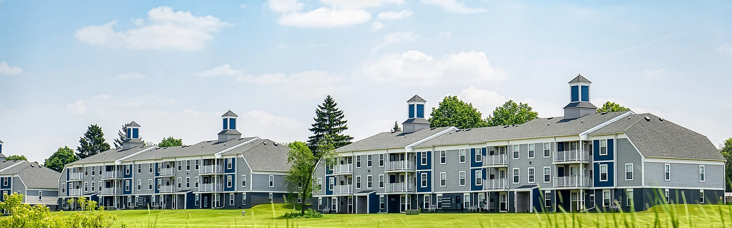 Elegant apartment complex surrounded by lush greenery and clear blue skies, featuring modern architecture with white and blue accents.