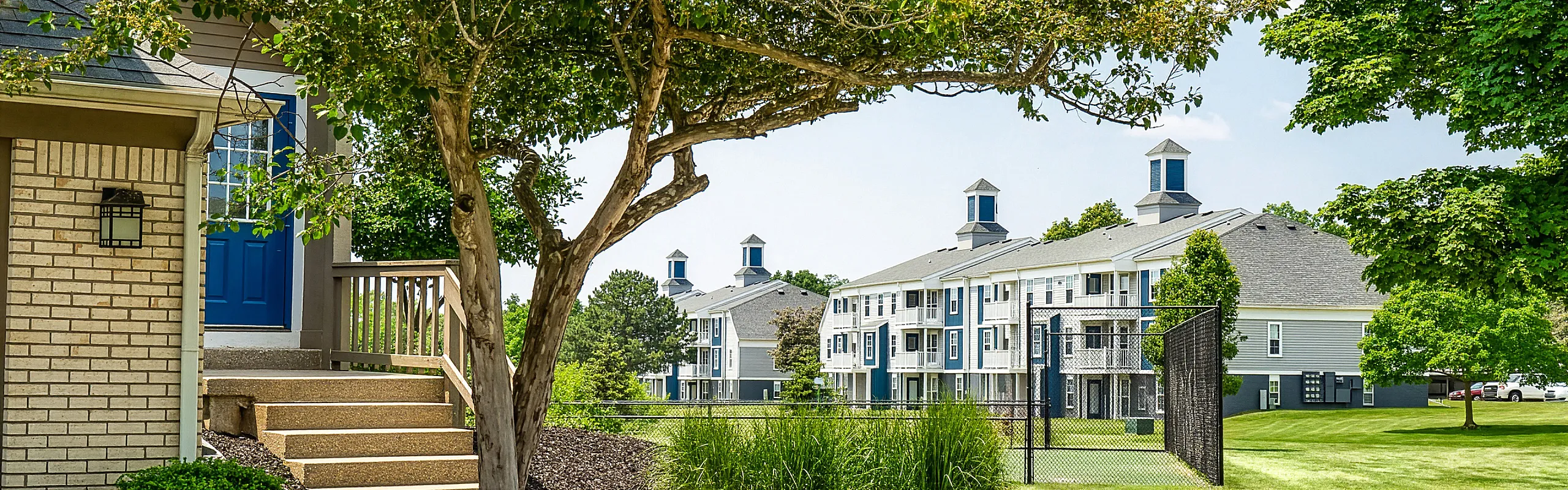 Charming residential complex with modern architecture, surrounded by lush greenery and sunlit pathways, featuring elegant blue and white buildings.