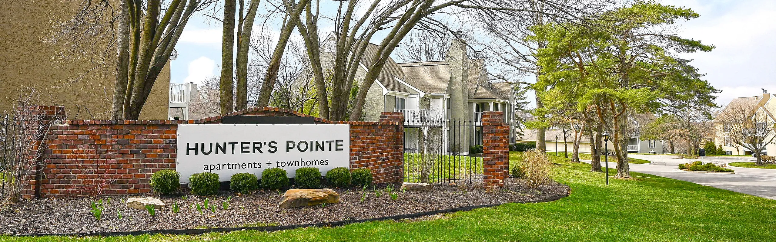 Welcoming entrance to Hunter's Pointe featuring lush greenery, charming brick accents, and elegant townhomes in a serene setting.