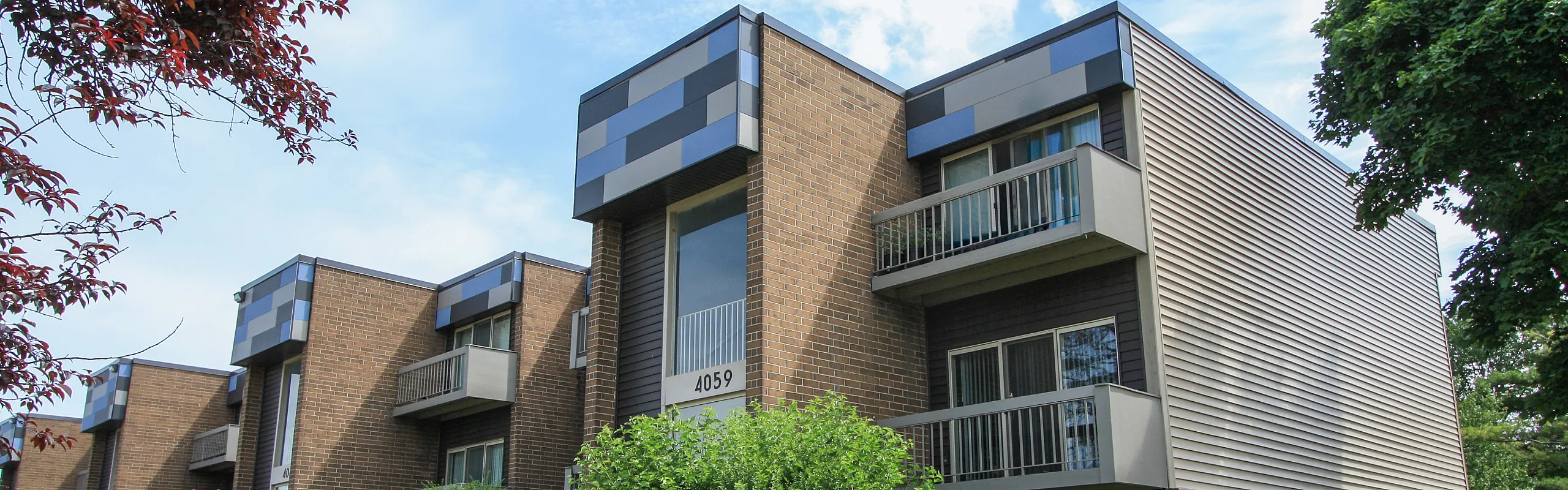 Modern apartment building with sleek brick facade and stylish balconies, surrounded by lush greenery and under a bright blue sky.