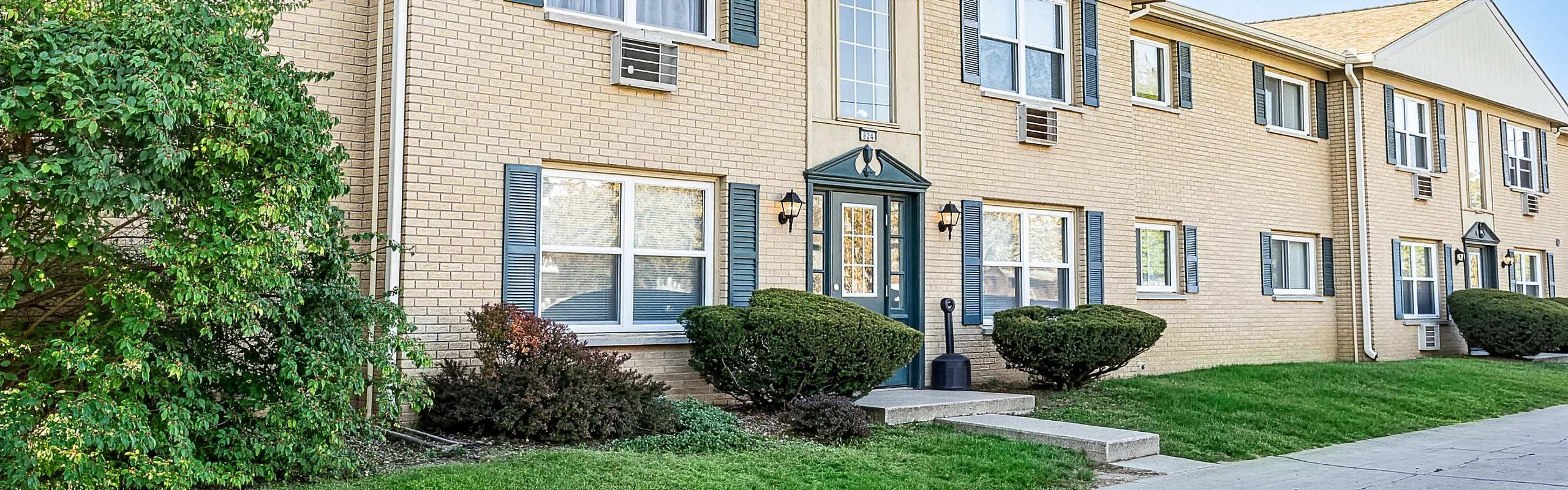 Charming brick apartment building with blue shutters and lush greenery, offering a welcoming and cozy atmosphere.