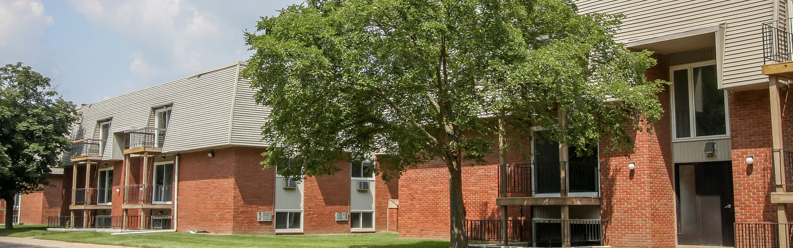 Charming brick apartment exterior with lush green landscaping and bright balconies, offering a serene and welcoming living space.
