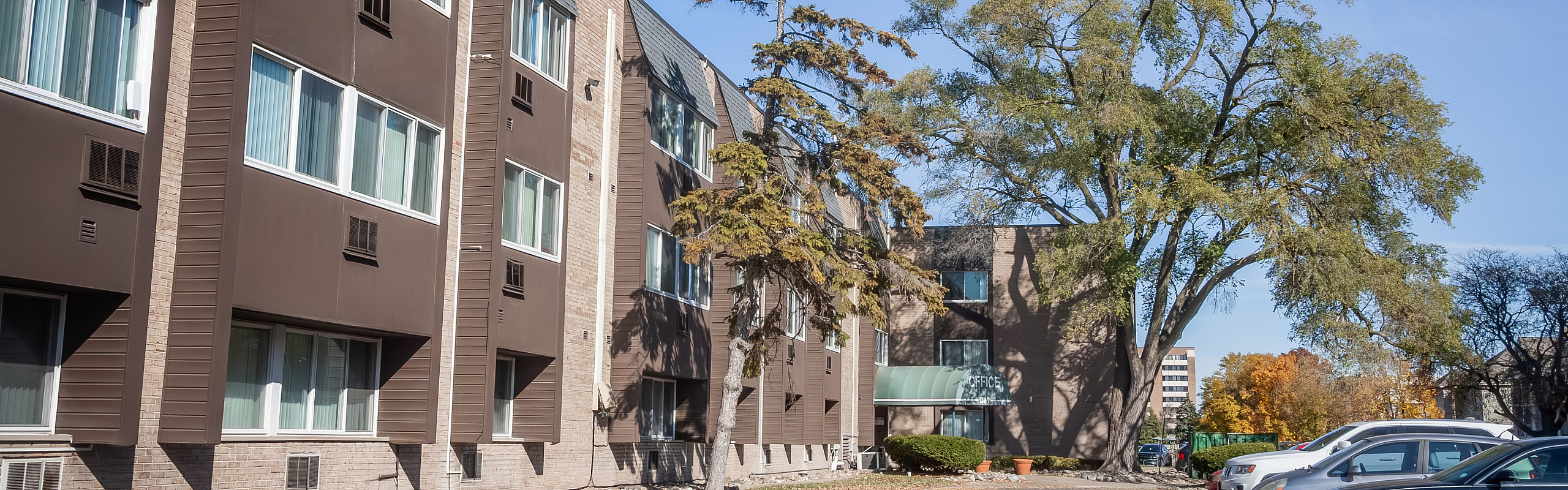 Charming apartment building with modern exterior, set amid lush trees and a sunny parking area, creating a welcoming urban living space.