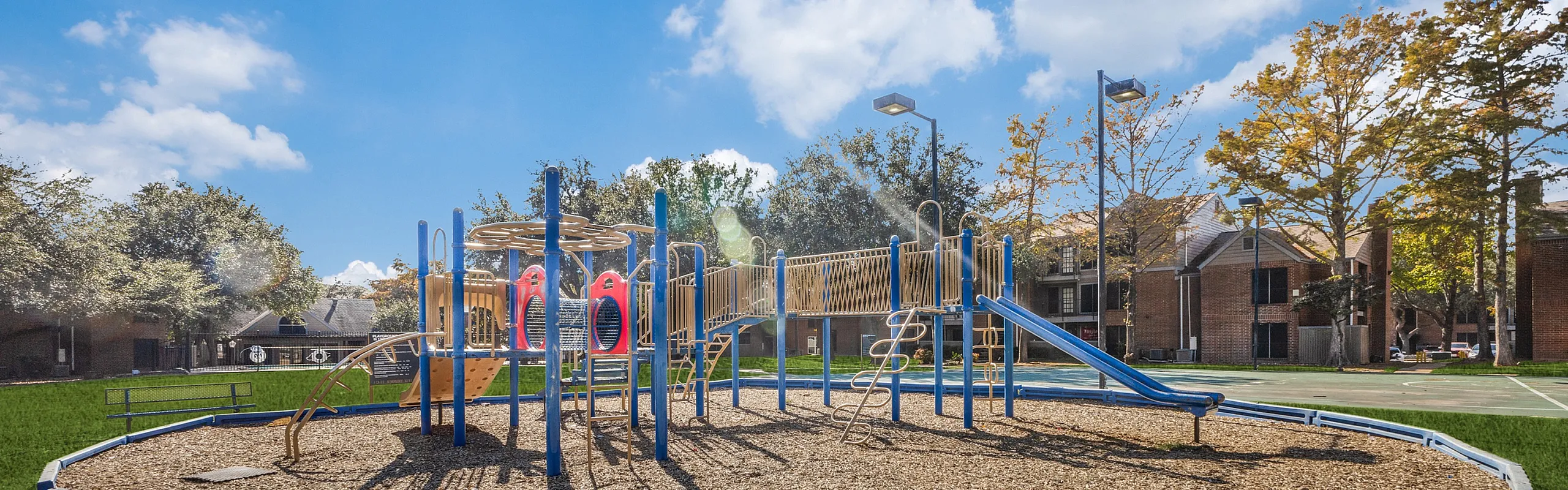 Charming apartment complex featuring a colorful playground set against a backdrop of lush greenery and clear skies, perfect for family-friendly living.