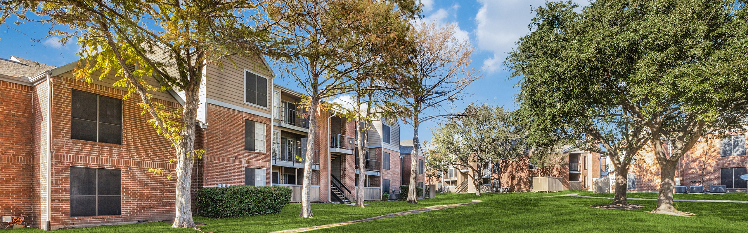 Charming red brick apartment buildings surrounded by lush greenery and mature trees under a clear blue sky.