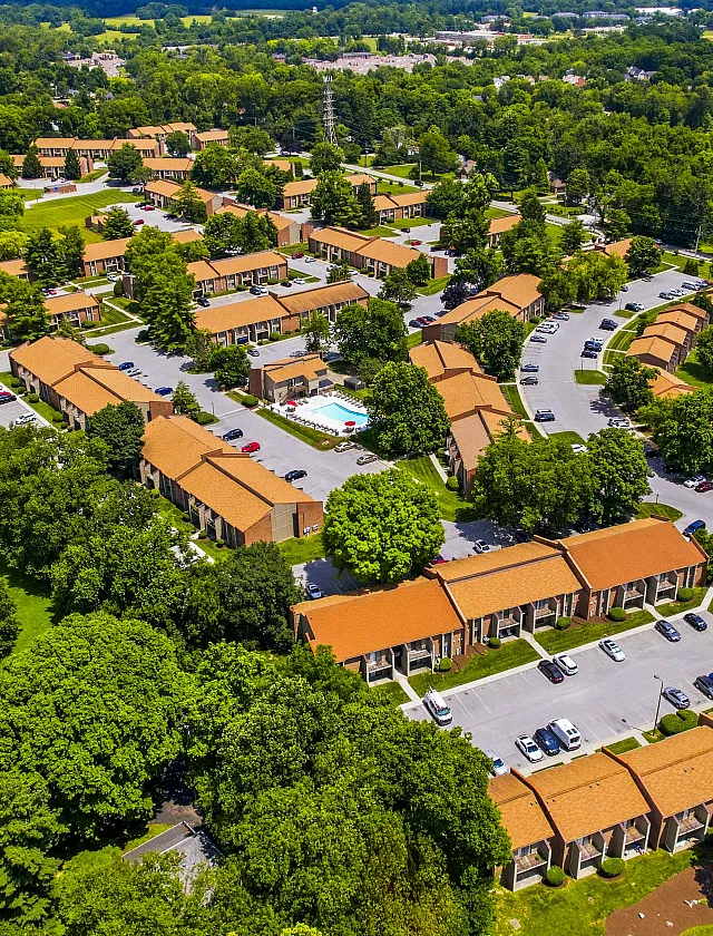 Aerial view of a charming apartment complex with red-roofed buildings nestled among lush greenery, featuring ample parking and a central swimming pool, creating an inviting and serene living environment.
