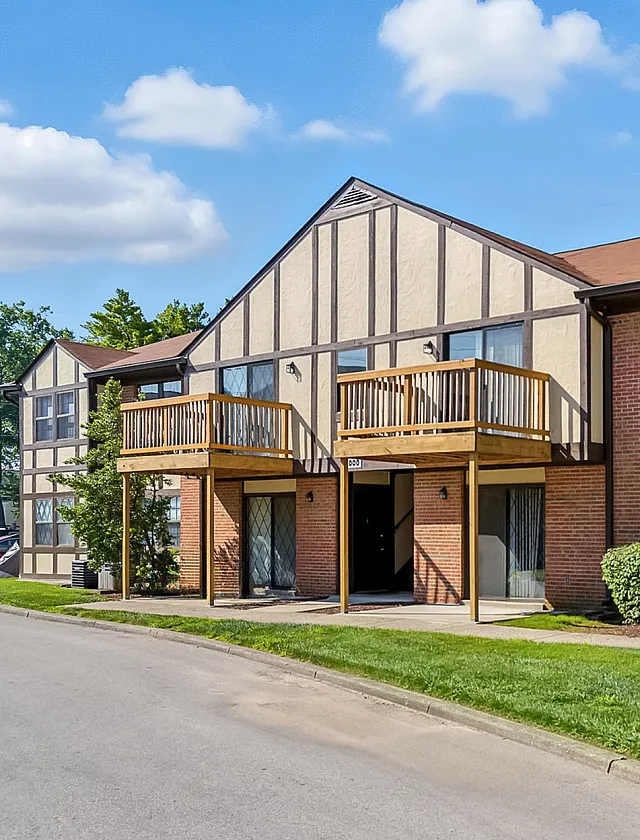 Charming two-story apartment building with wooden balconies, featuring Tudor-style architecture and surrounded by lush greenery, under a bright blue sky.