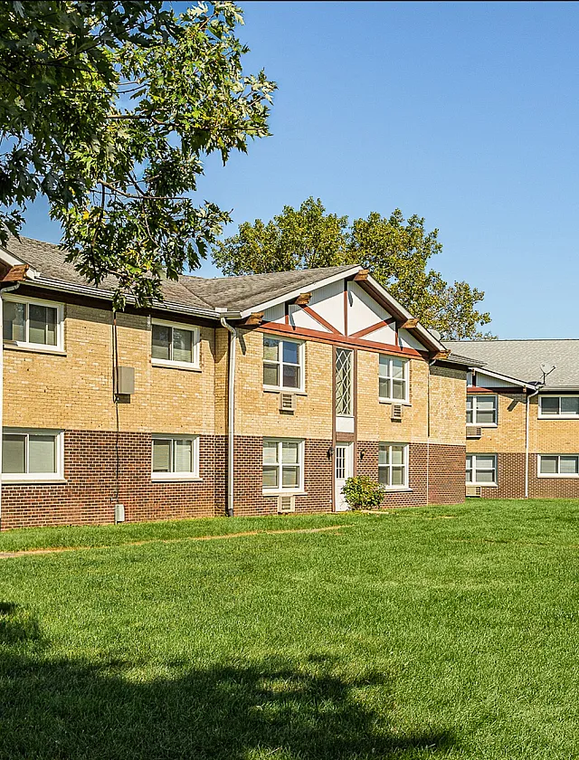 Charming brick apartment complex with manicured lawns and lush trees under a clear blue sky.