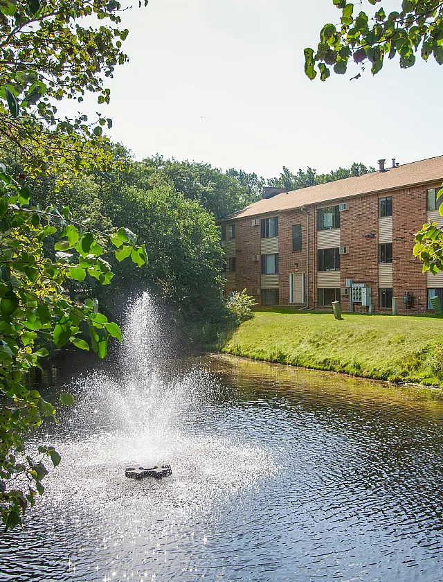 Charming apartment building nestled by a serene pond with a sparkling fountain, surrounded by lush greenery.