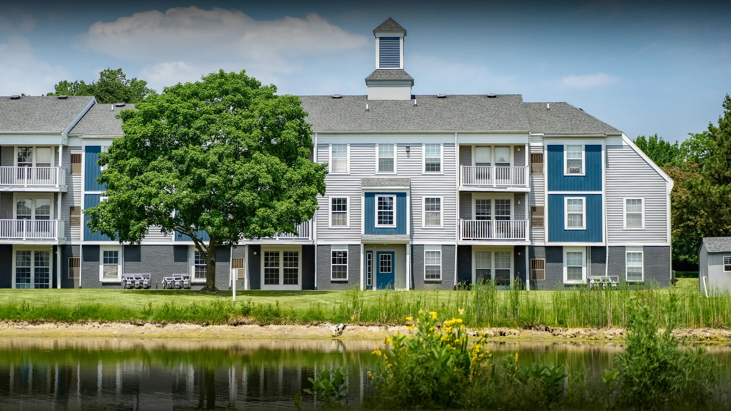 Charming apartment complex with modern blue and white siding, lush green surroundings, and serene pond view.