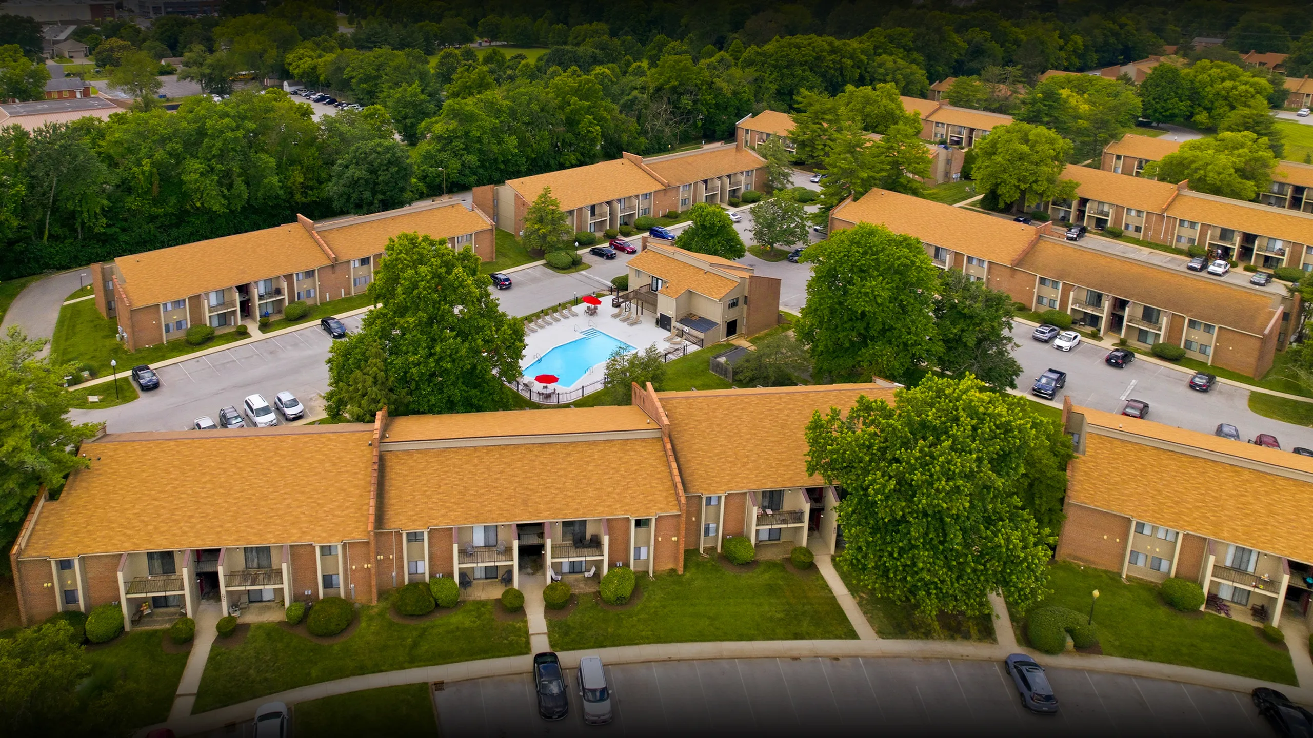 Scenic aerial view of a charming apartment complex surrounded by lush greenery, featuring a sparkling central pool area and ample parking space.