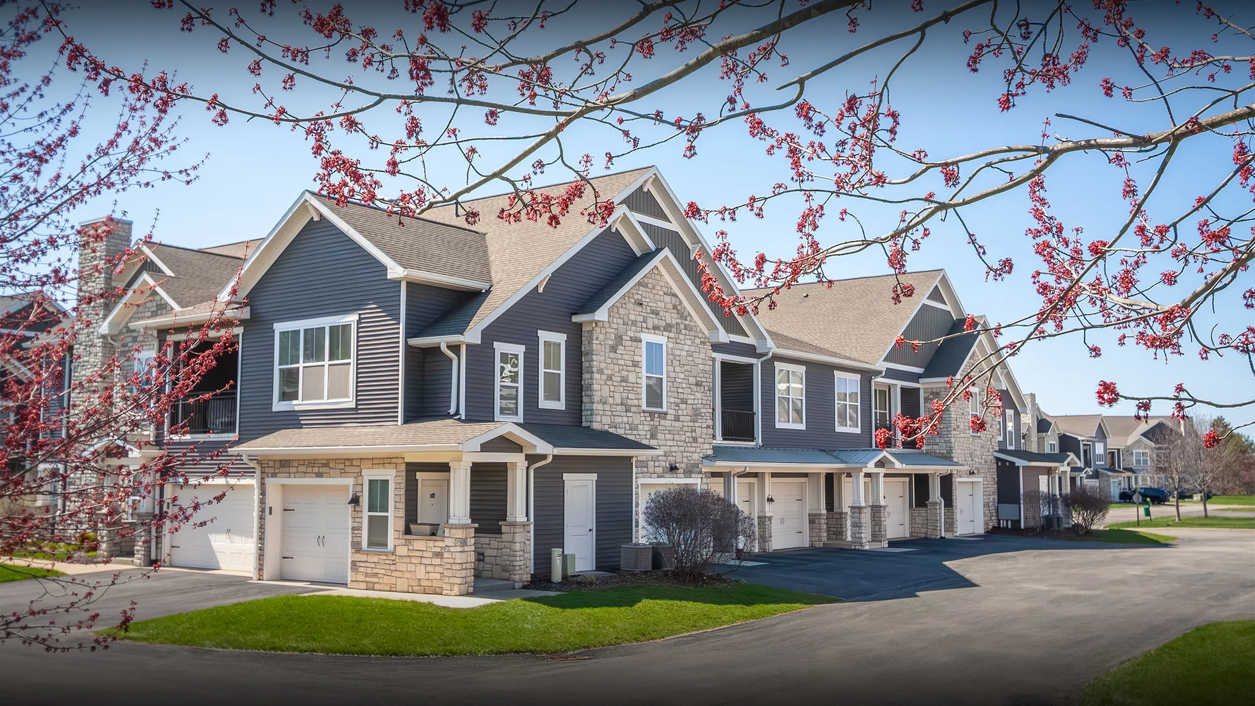 Charming modern townhouses with stone and siding facade, surrounded by blossoming trees and well-maintained lawns.