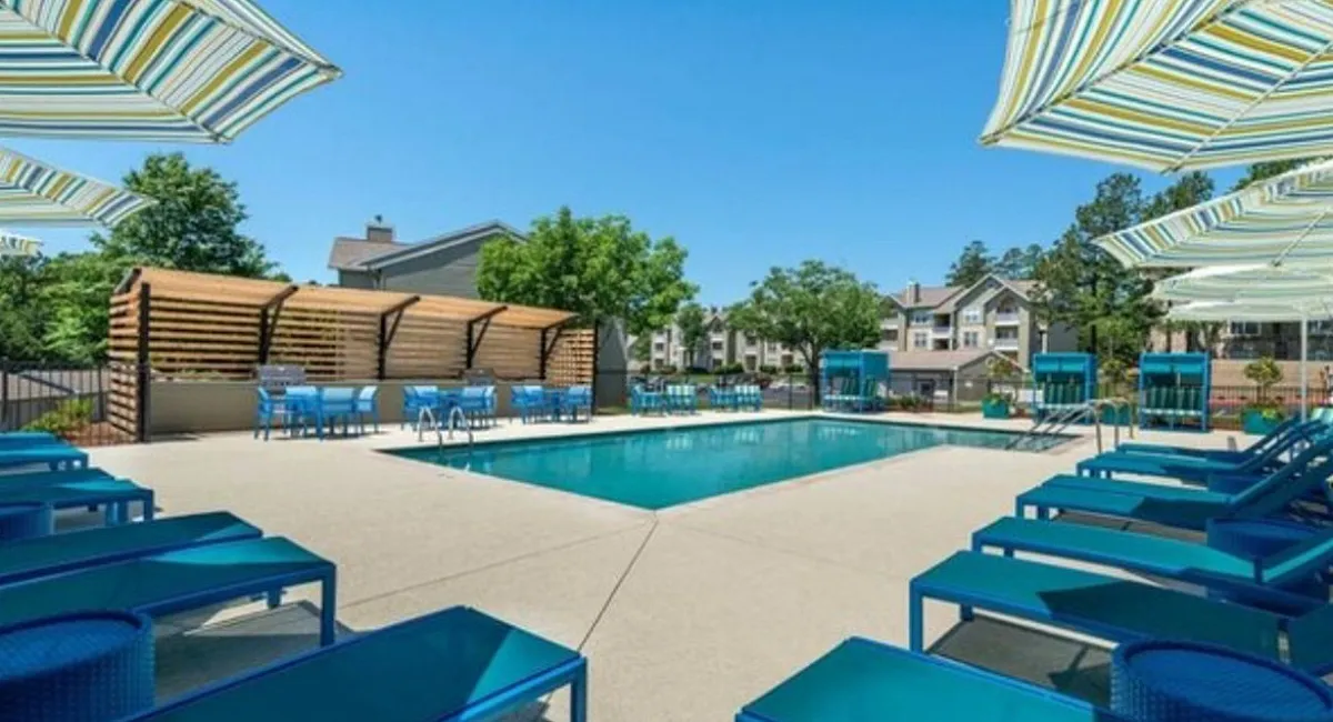 Luxurious apartment complex featuring a vibrant outdoor pool area with chic lounge chairs and elegant striped umbrellas, surrounded by lush greenery and stylish architecture.