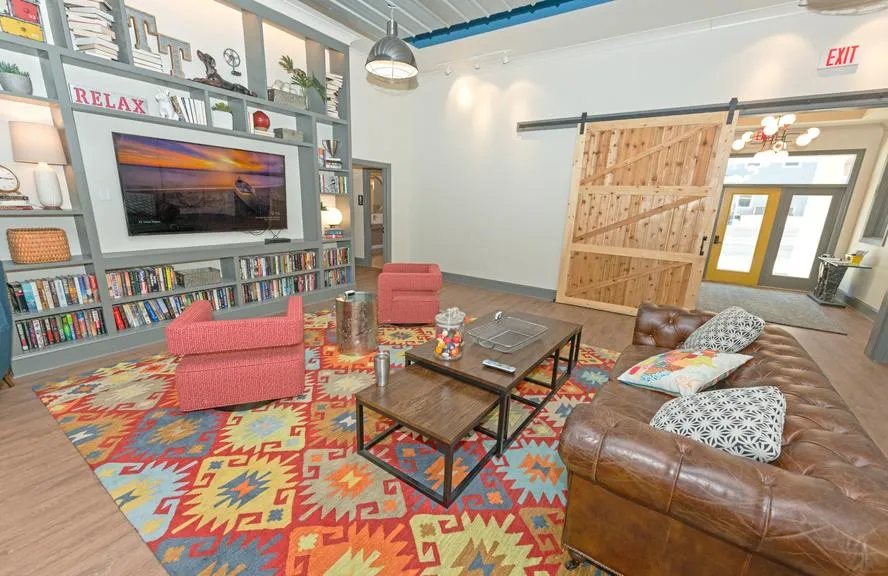 Chic communal lounge with cozy red chairs, leather sofa, vibrant patterned rug, and a TV on a stylish bookshelf wall, perfect for relaxation.
