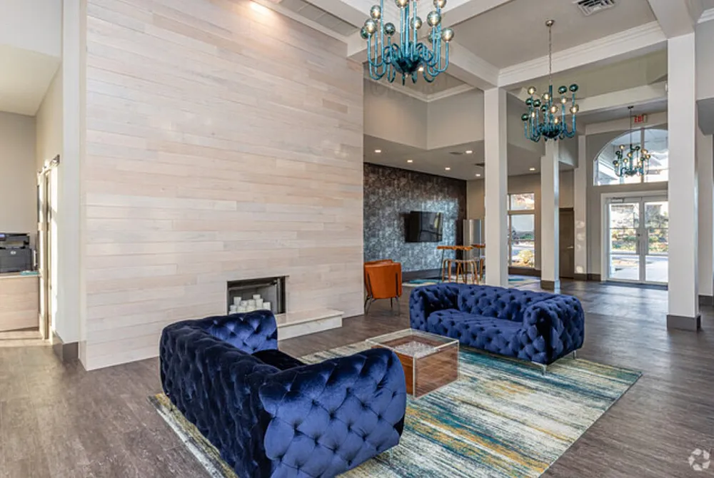 Elegant apartment lobby featuring plush blue velvet sofas, modern chandeliers, and a cozy fireplace with ample natural light.