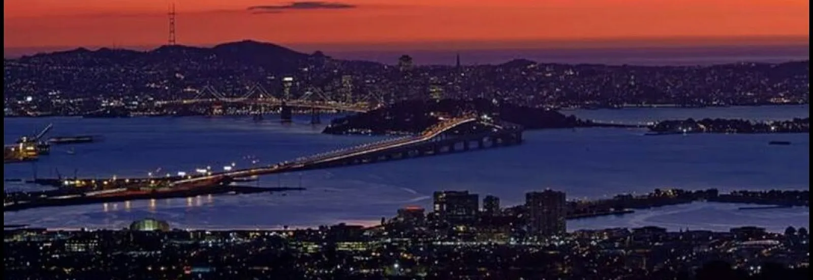 Breathtaking panorama of a vibrant sunset over a sprawling cityscape with a beautifully lit bridge connecting the skyline across calm waters.