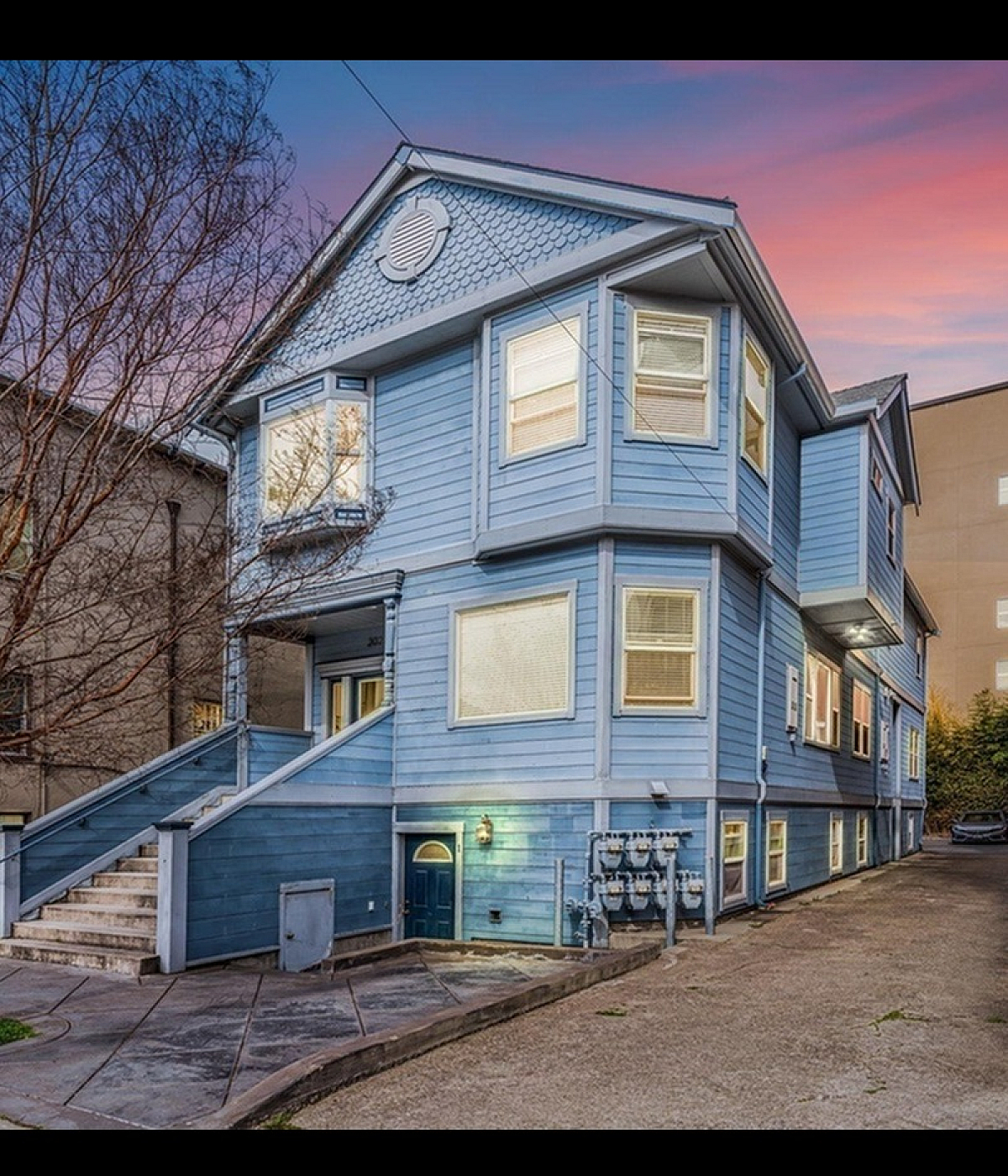 Charming blue multi-story apartment building with bay windows, nestled in a serene urban setting during a vibrant sunset.
