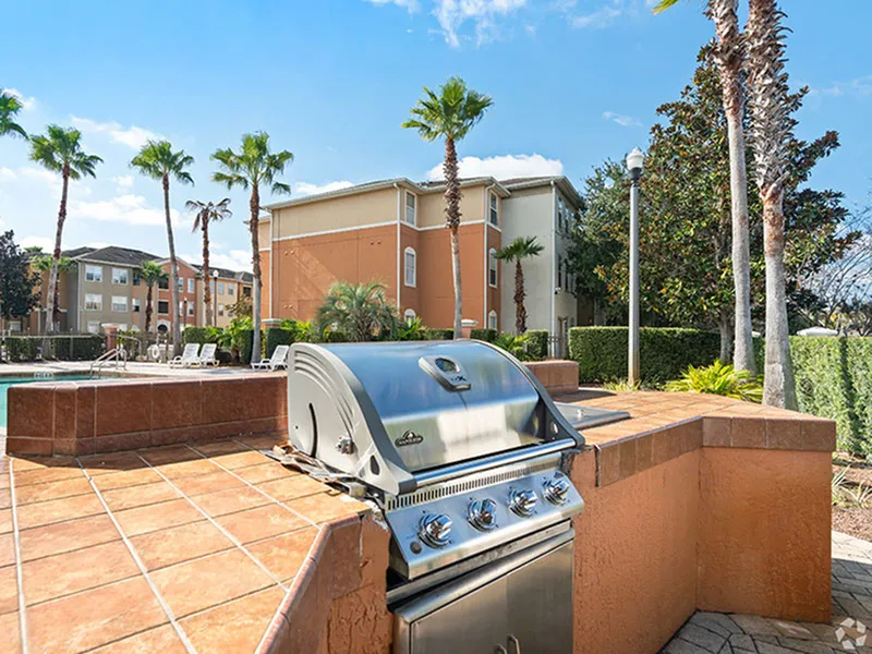 Luxurious outdoor grilling area with modern BBQ set amid palm trees and a vibrant apartment community backdrop, perfect for entertaining.