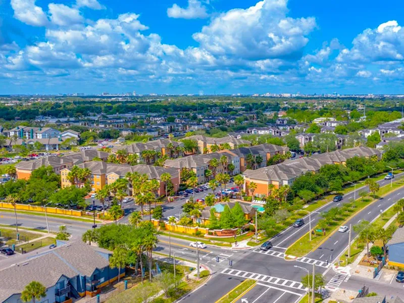 Vibrant aerial view of a beautifully landscaped apartment complex surrounded by lush greenery and clear blue skies, offering a serene and inviting living environment.