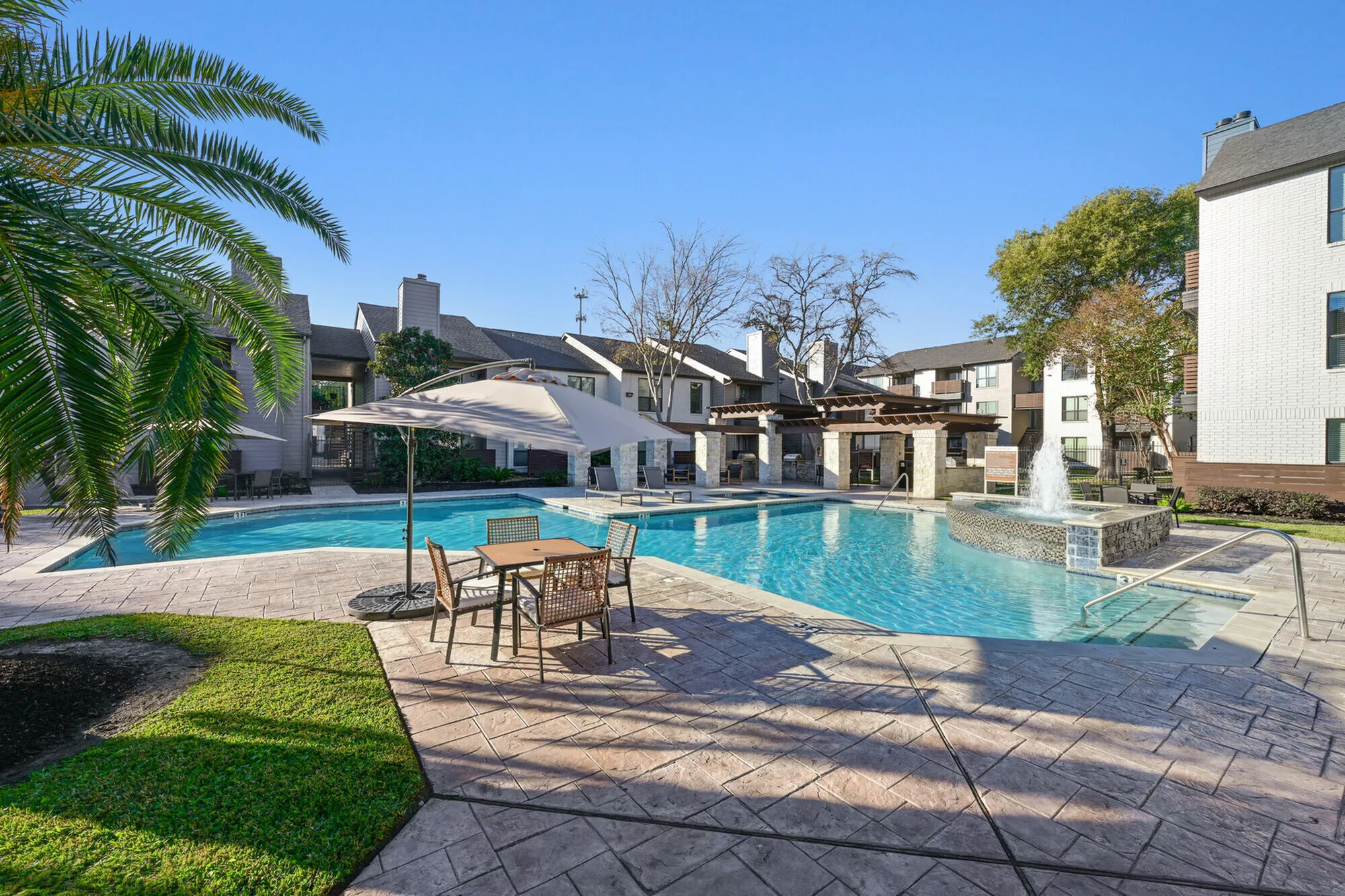 Luxurious apartment community pool area with sparkling fountain, modern umbrella-shaded seating, and lush greenery, perfect for relaxation and leisure.