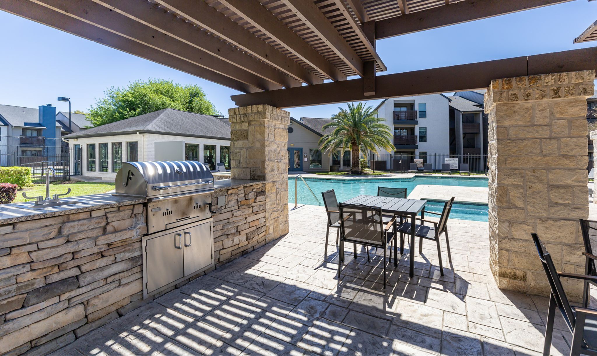 Inviting outdoor patio with a built-in grill, overlooking a sparkling pool and palm trees at a modern apartment complex.