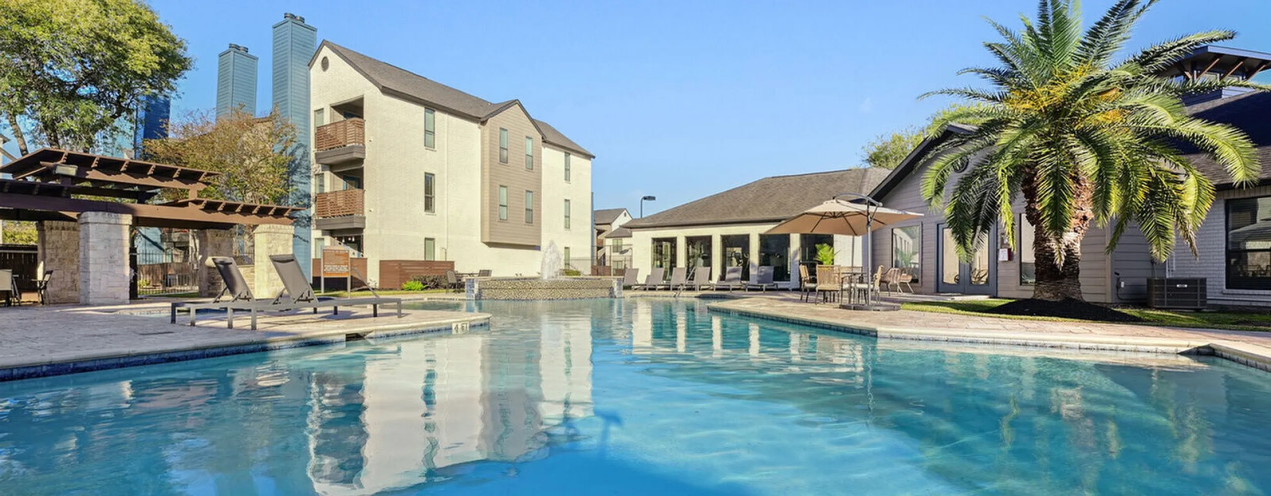 Luxurious apartment complex with a sparkling pool, palm trees, and elegant outdoor lounge area under a clear blue sky.