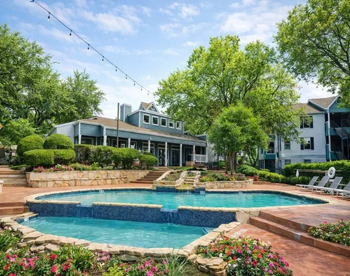 Inviting apartment complex with beautifully landscaped gardens, a sparkling multi-level pool, and charming architecture under a bright blue sky.