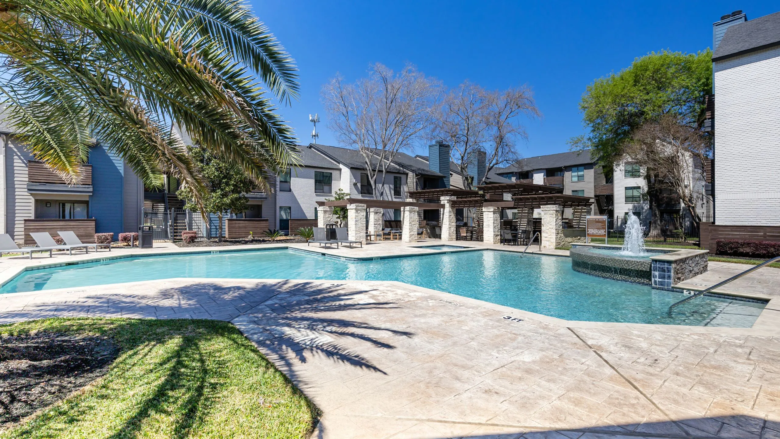 Luxurious apartment complex featuring a sparkling pool with fountain, surrounded by modern buildings and lush palm trees.