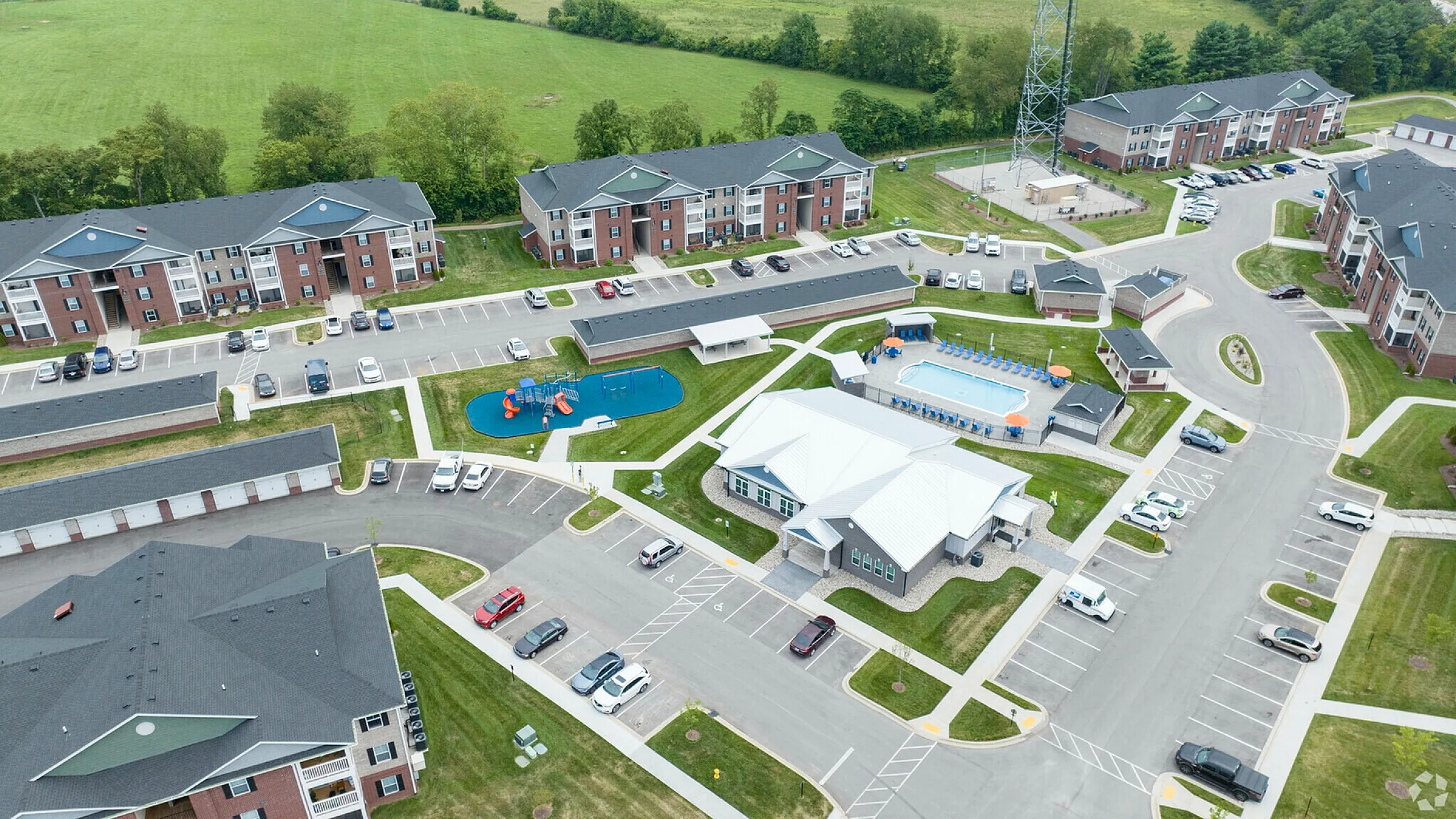 Aerial view of a vibrant apartment community featuring modern brick buildings, ample parking, a sparkling swimming pool, playground, and lush green surroundings.