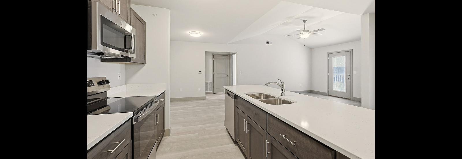 Modern open concept kitchen with sleek stainless steel appliances, double sink, and stylish cabinetry leading to a bright living area with ceiling fan and balcony access.