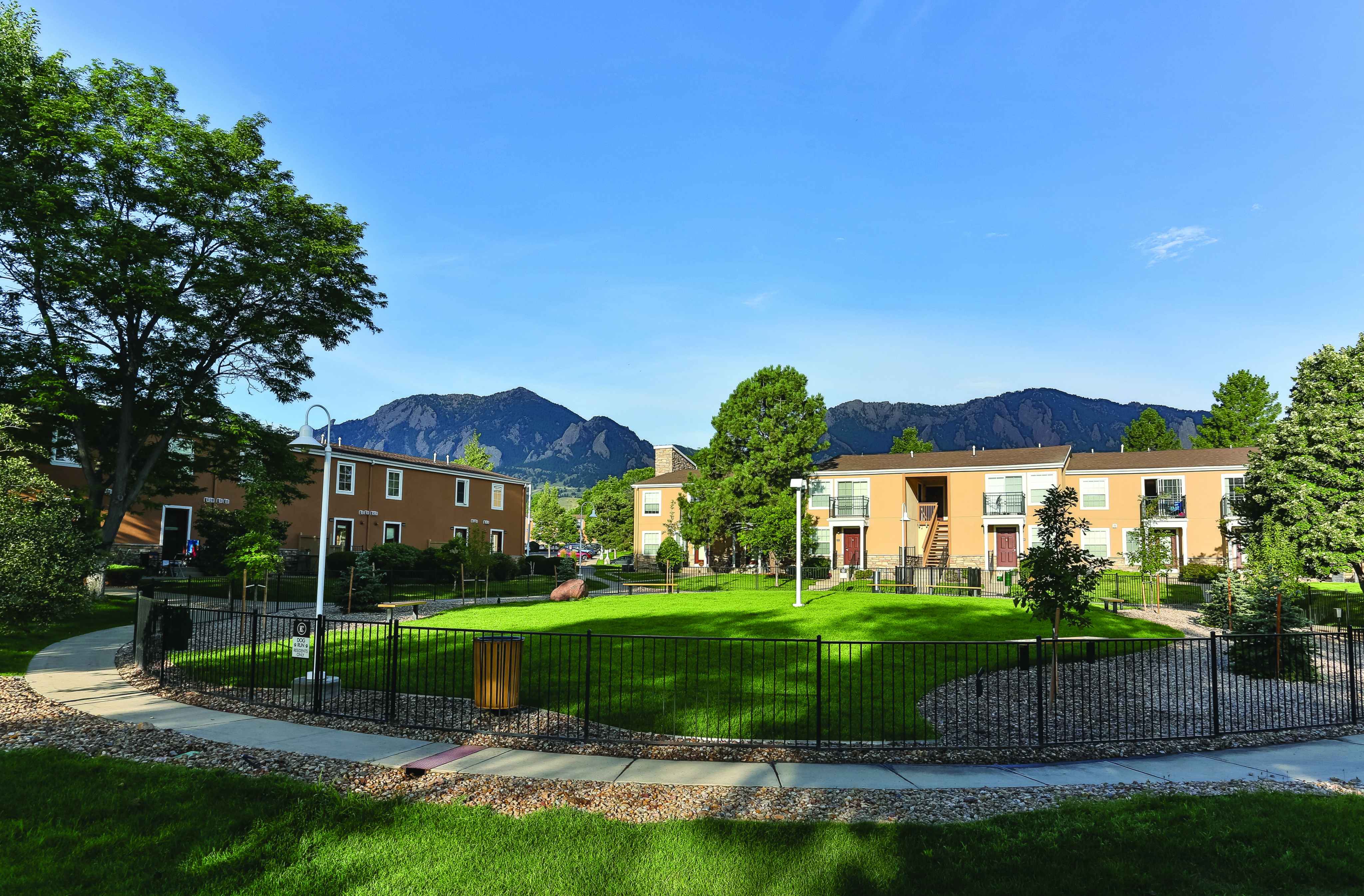 Charming apartment complex with lush green lawns, surrounded by scenic mountain views under a bright blue sky.